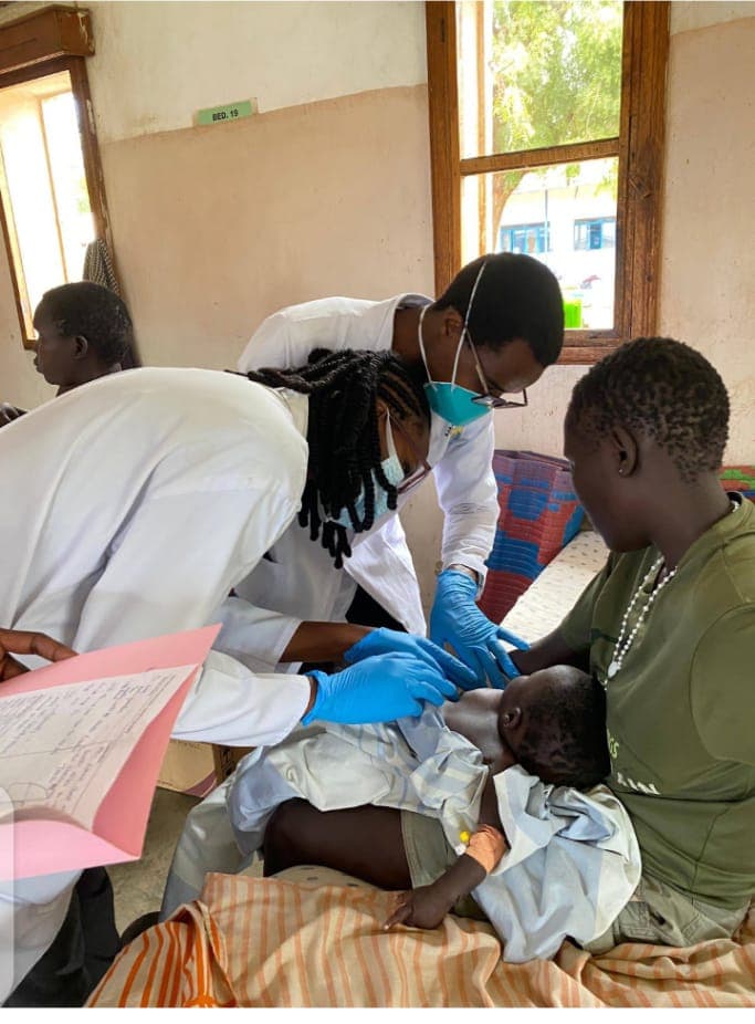 Three clinicians in white coats, one wearing surgical gloves, examining a small child held by a woman on a hospital ward bed.