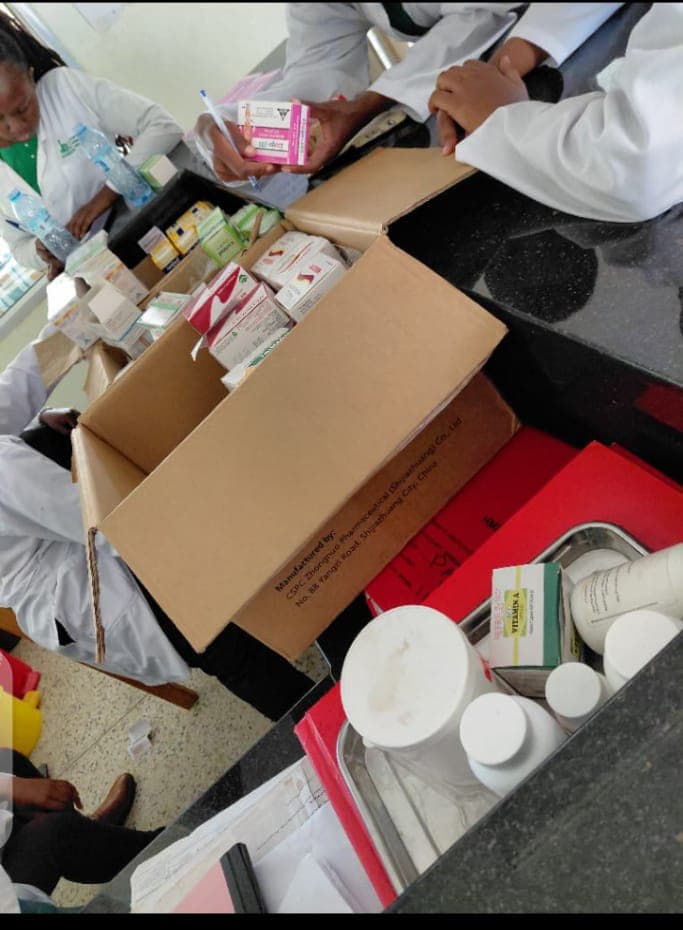 Health workers in white coats standing around a counter, unpacking boxed medicines from a cardboard carton and writing down what is inside.