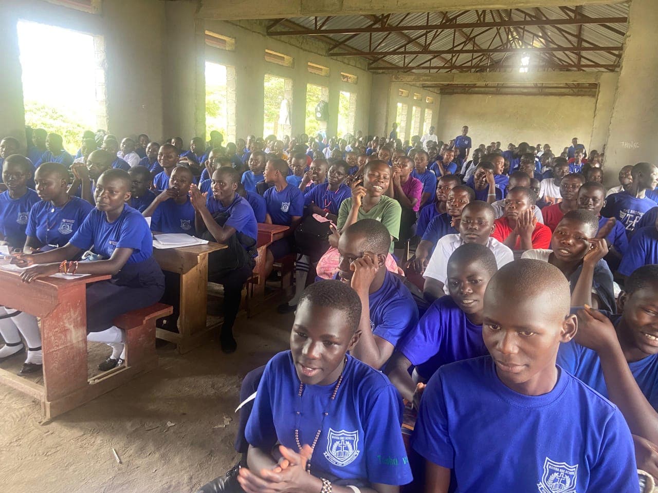 A hall packed with secondary school students in blue uniforms, seated at desks and on the floor, listening during a health talk.